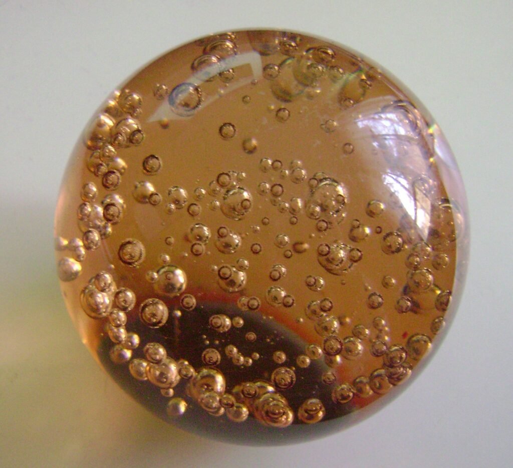 A domed amber glass paperweight seen from above, filled with a rising trail of round air bubbles of varying size.