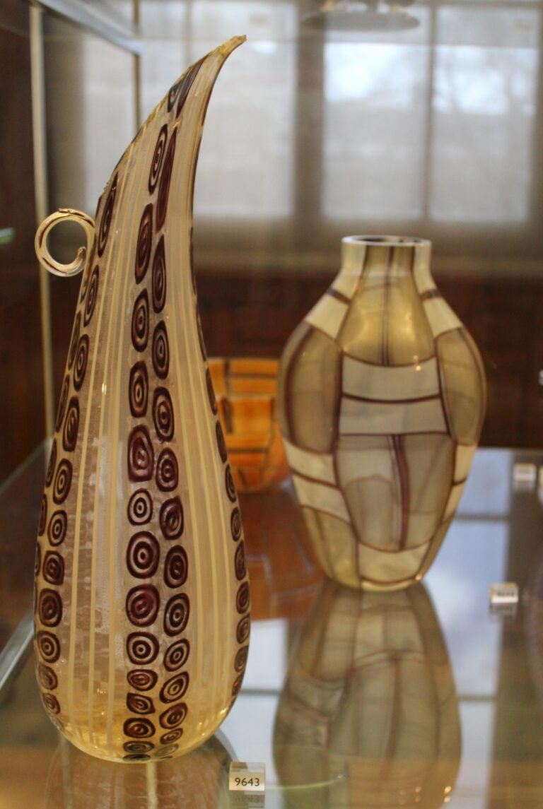 Two Murano glass vases in a museum case: a tall leaf-shaped amber vase patterned with dark spiral murrines, and behind it a rounded vase in a pale grid pattern.