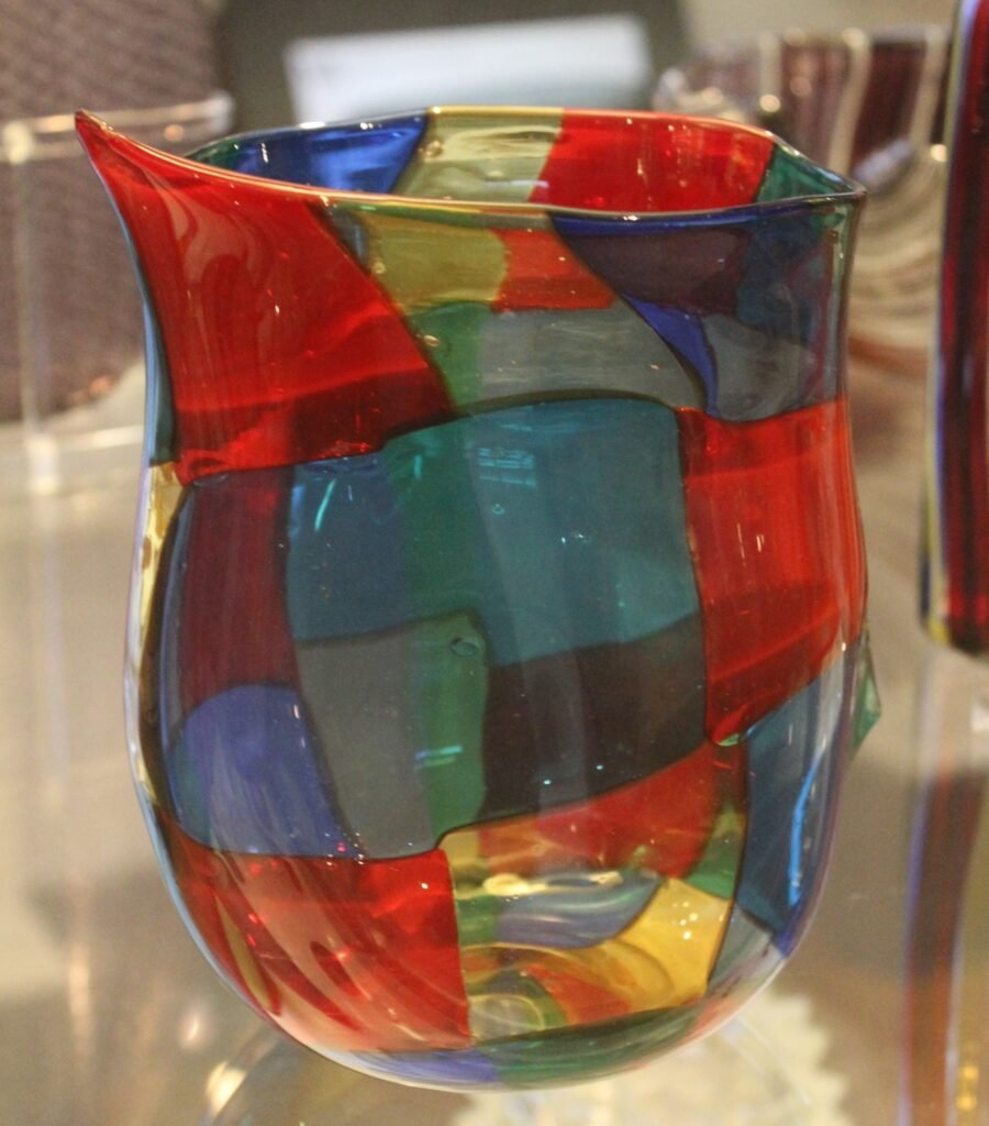 A blown glass vase made from irregular squares of red, blue, green, yellow and clear glass fused together, with an asymmetric flared rim.