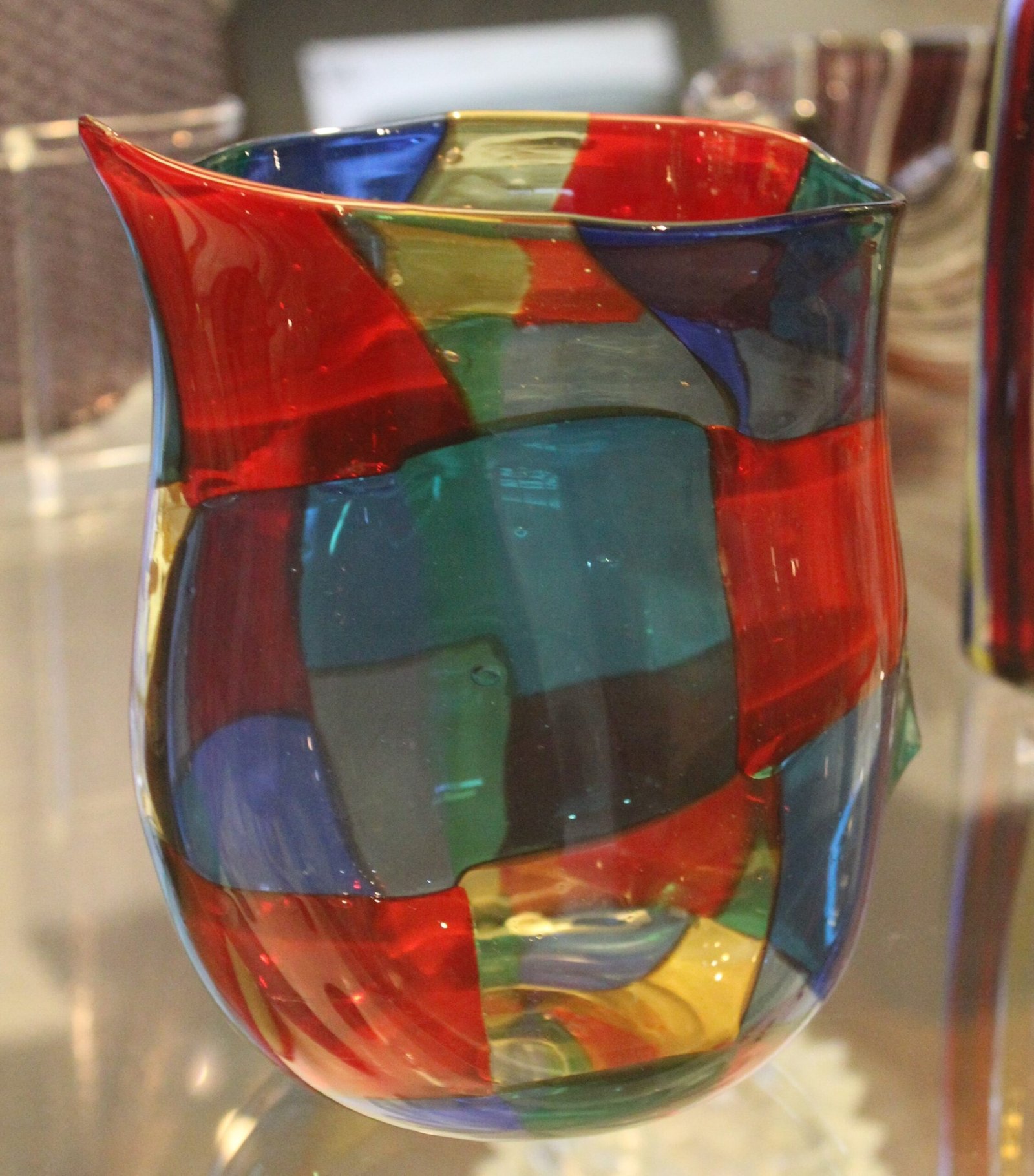 A blown glass vase made from irregular squares of red, blue, green, yellow and clear glass fused together, with an asymmetric flared rim.