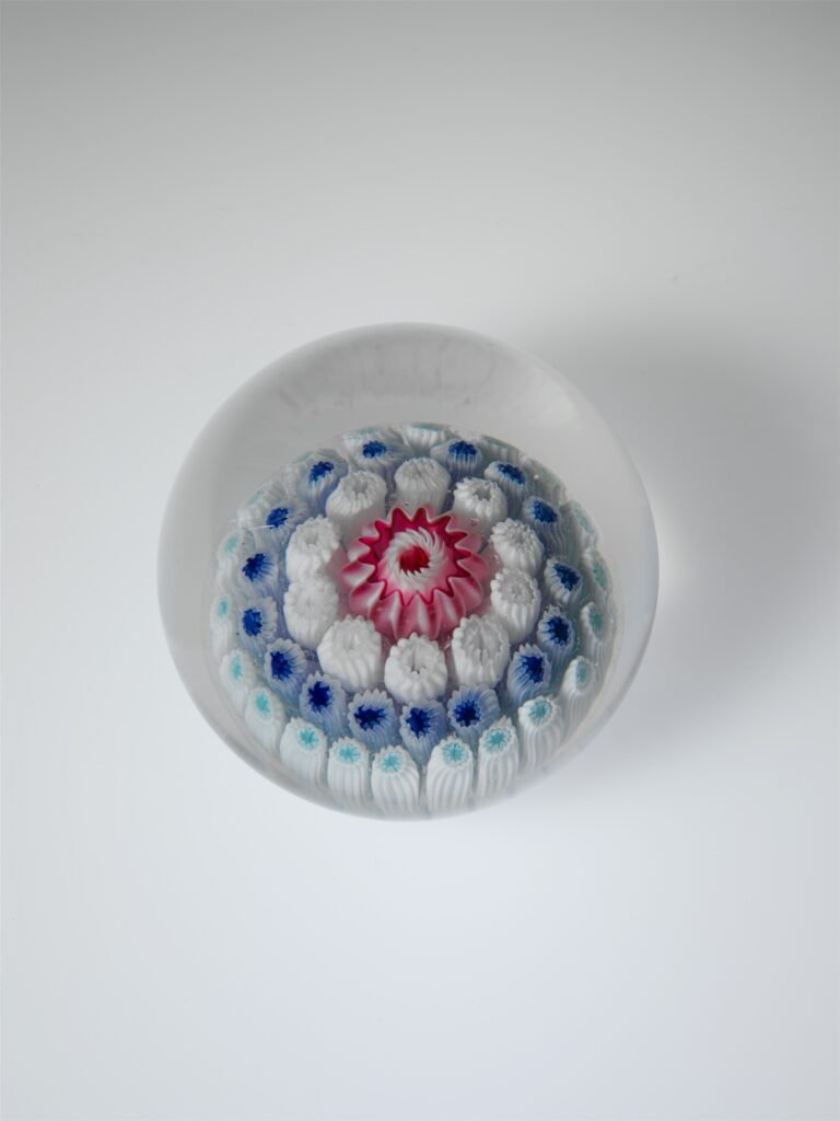 A domed glass paperweight seen from above, with a red and white central floret ringed by concentric rows of blue and white millefiori canes.