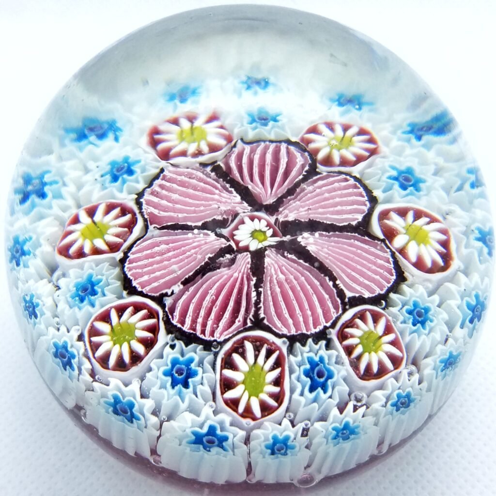 A millefiori paperweight seen from above: a pink and white flower at the centre, ringed by rows of small blue and white canes.