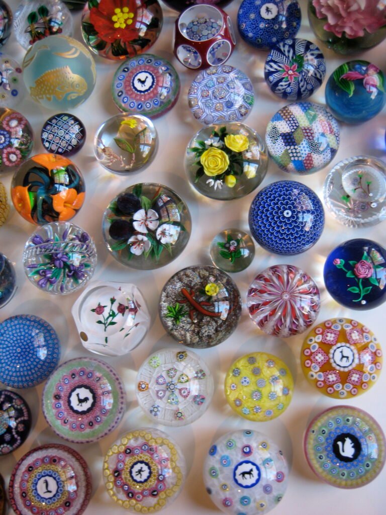 Several dozen glass paperweights arranged together in rows, showing a wide range of millefiori, floral and abstract designs.