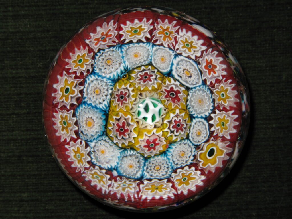 A close-packed millefiori paperweight seen from directly above, with concentric rings of white, yellow and red florets on a deep red ground.