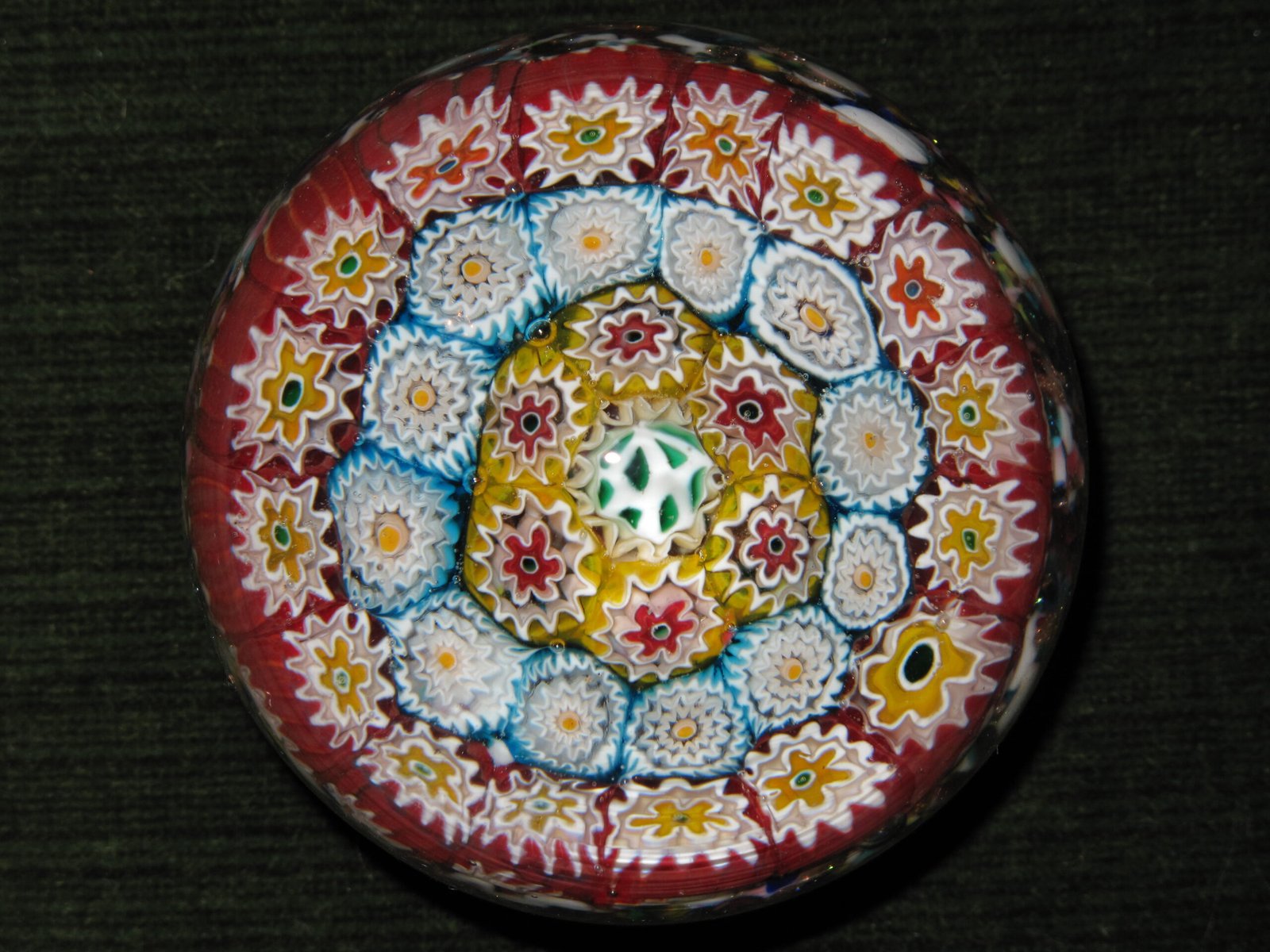 A close-packed millefiori paperweight seen from directly above, with concentric rings of white, yellow and red florets on a deep red ground.